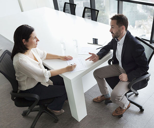A person seated across a table from another individual in a modern office setting, engaged in a discussion while reviewing documents.